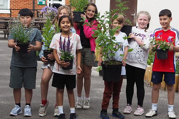 Seven primary school age children in outdoor clothes standing in a playground and holding a variety of small potted plants