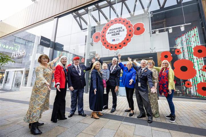 Ten people standing in a horse shoe shape and pointing to a shop that is decorated in giant red poppies.
