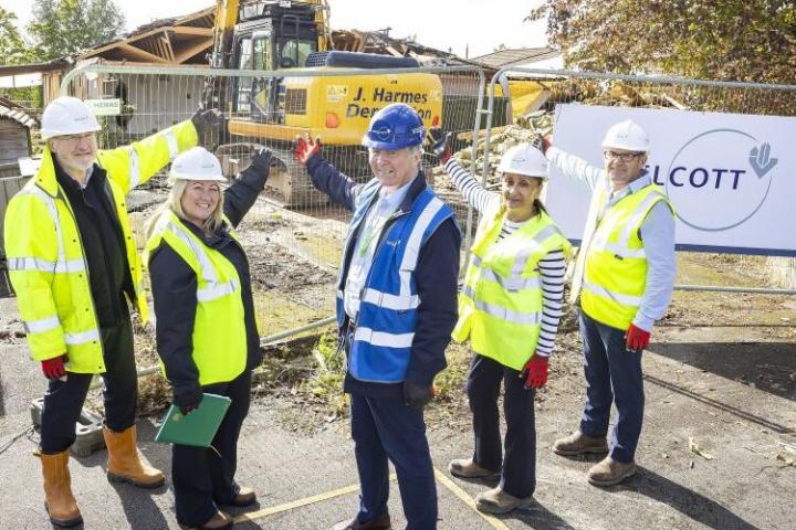 Five people in high-vis jackets and hard hats standing in a line a pointing at a digger