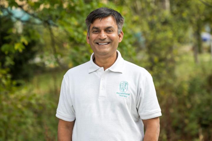 A man with dark hear wearing a white polo shirt with a green occupational therapy logo smiles at the camera.