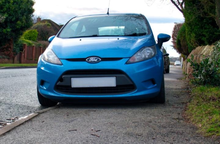 Blue car parked half way up a pavement on a residential street with shrubs, trees and fences in the background