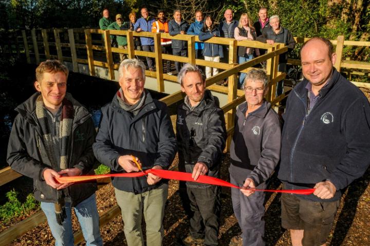 5 members of the bridge replacement project team standing in a line in front of the bridge. They are holding a red ribbon and one person is holding a pair of scissors.