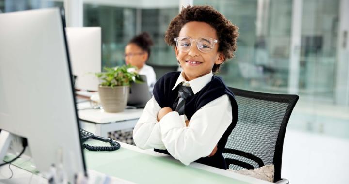 A child sitting at an office desk, facing a computer monitor, with another child working in the background.
