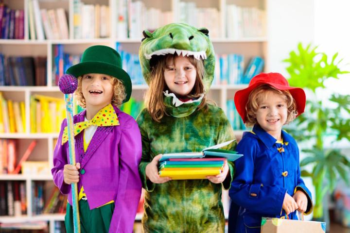 Three children wear fancy dress in front of colourful bookshelves. They are dressed as Willy Wonka, a dinosaur, and Paddington Bear.
