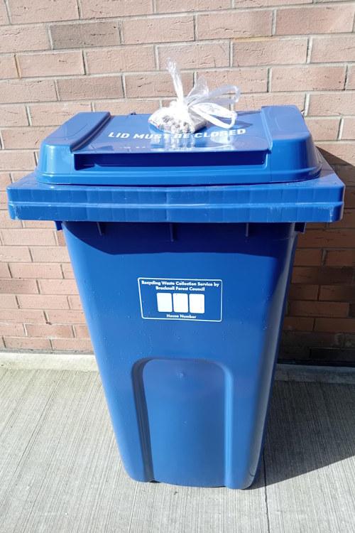 A blue recycling bin with a tied-up, clear plastic bag full of household batteries on the closed lid.