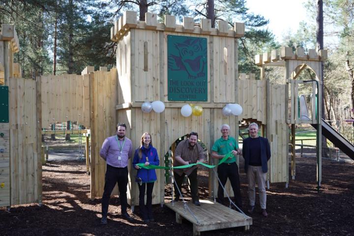 A group of people stand within the new fort at the Look Out, cutting a green ribbon. The fort is a large, wooden construction shaped like a castle.