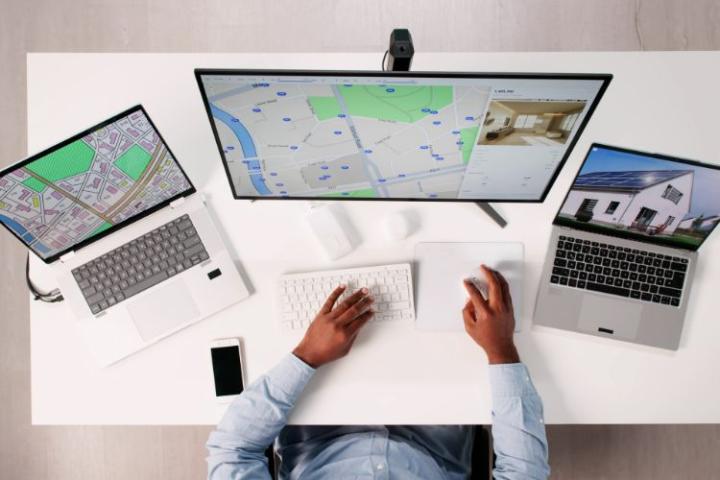 A man in a blue shirt at a computer that has maps on the screen. A laptop is either side of the main screen and have property images on them. 