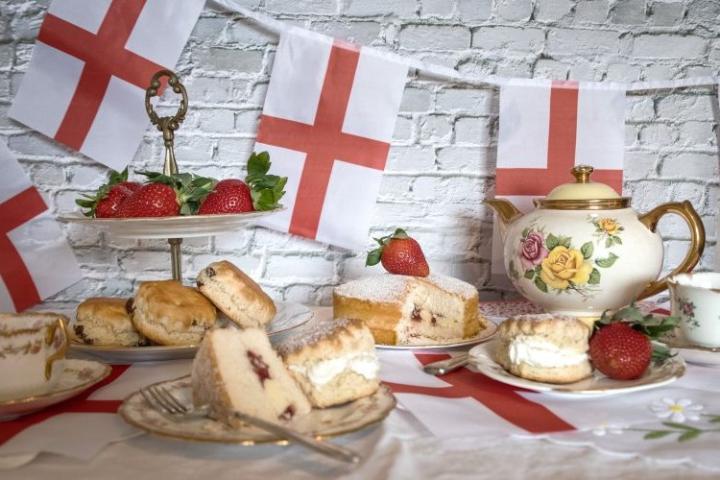 Tea party setup with English flag bunting on a brick wall. A floral teapot and teacups are on a table with scones, strawberry-topped sponge cake, and fresh strawberries. Plates and a tiered stand hold these treats, creating a festive atmosphere.