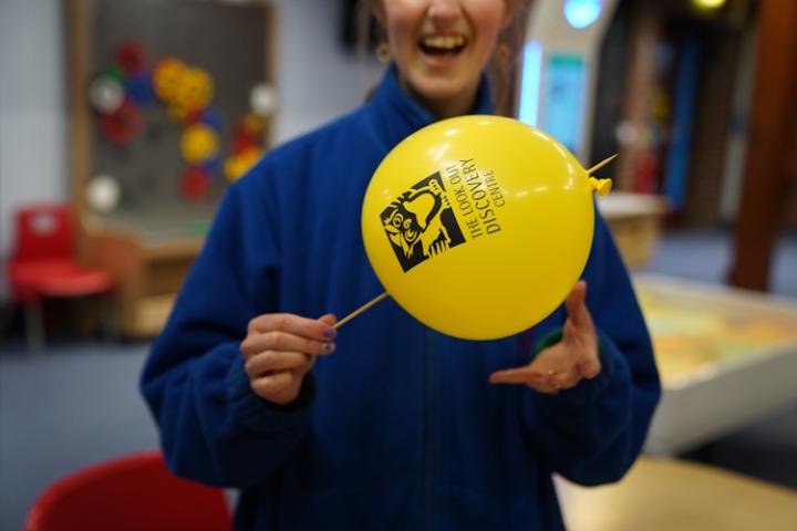 A person in a blue fleece sticks a pointed wooden skewer through a bright yellow balloon without popping it.