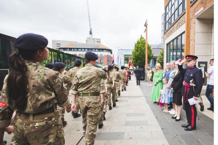 A parade of people in military uniforms marching through Bracknell town centre being observed and saluted by bystanders 
