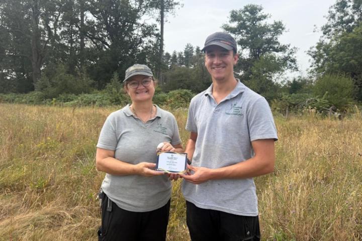 Two people stand in Buckler's Forest. They are holding the Land Trust's 2026 Site of the Year award.