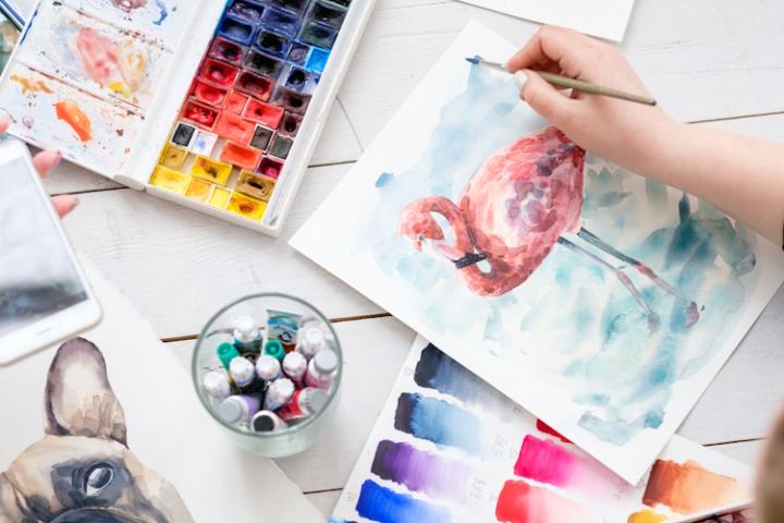 A person paints a watercolour of a flamingo on a blue background. Beside them are several other watercolours, including test pages and a painting of a dog.