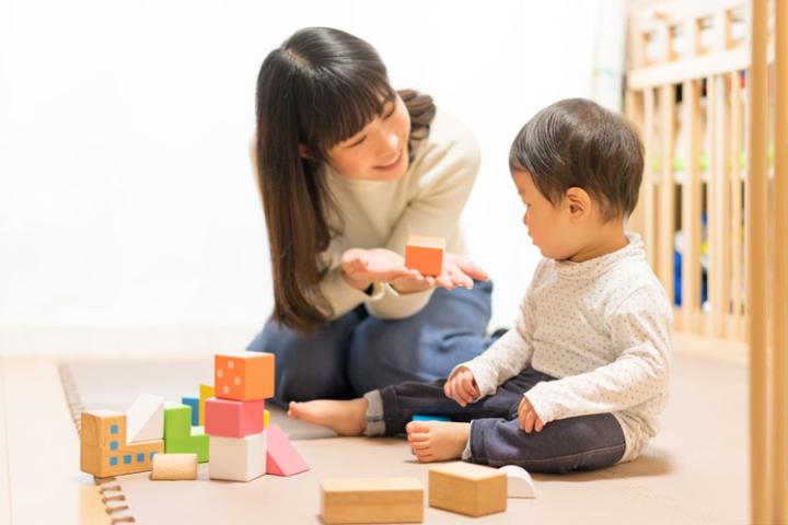 An adult sitting on a floor mat with a young child, using colourful wooden blocks during a play activity.