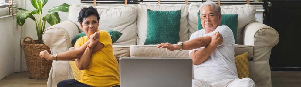 Two older people sat on the floor in front of a sofa, looking at a computer screen and doing arm stretches
