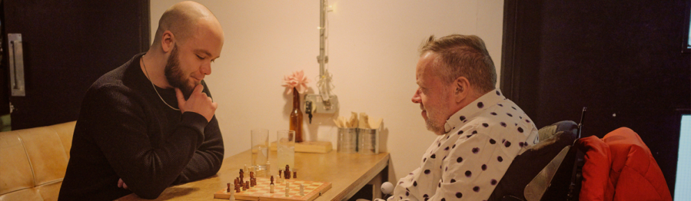 Two people play chess at a wooden table; the person on the right, in a wheelchair, wears a white shirt with black polka dots.