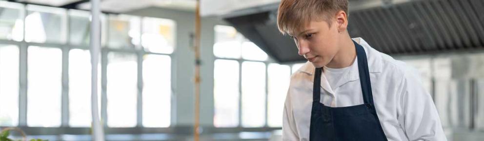 Young person in commercial kitchen wearing white overalls and blue apron