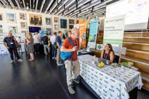 A busy room with stalls at Bracknell Forest Climate Summit 2025. A man speaks to a woman at a stall for helping homeowners get solar power.