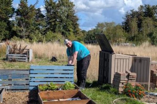 A man in a bright blue t-shirt and dark trousers leans over a blue pallet in an allotment. Behind him is a storage box and several piles of bricks. He is grinning, and it's a sunny day.
