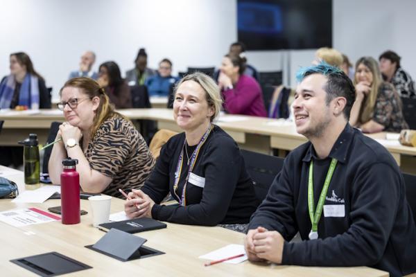 Three people sit at a desk at a busy conference, listening intently with smiles on their faces. 