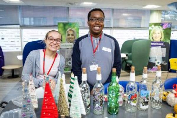 A man and a woman stand at a table covered in Christmas crafts, including decorated wool Christmas trees, sparkling lights in decorated bottles and knitted pumpkins. 