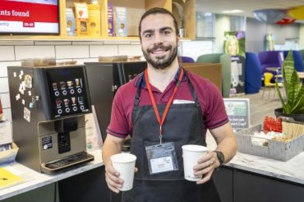 A smiling man stands beside a coffee machine with two cups of coffee. He wears a lanyard and apron.