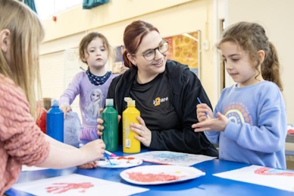 Three small children sit with an adult doing crafts. They are painting and decorating paper plates.