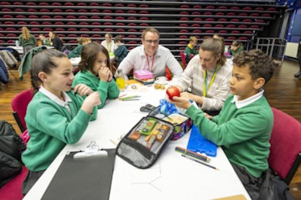 Children and two adults sit around a table looking at the contents of a packed lunchbox.