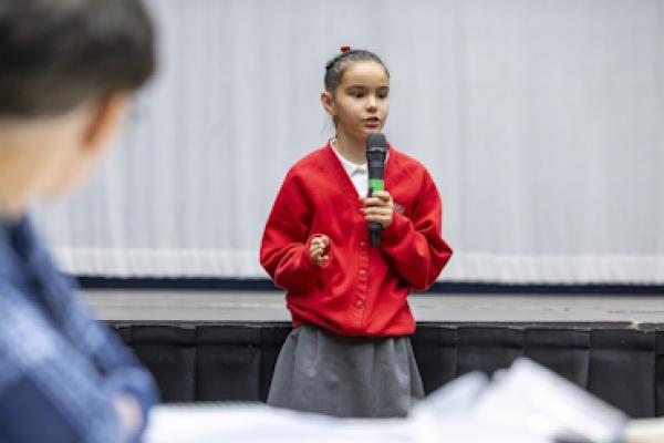 A primary-school aged girl in a red jumper stands and speaks into a microphone.