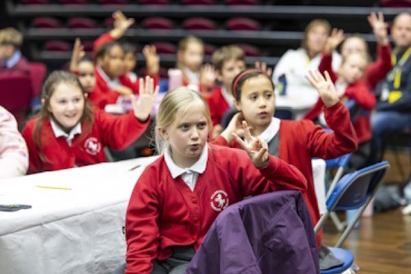 A group of children sat at tables holding up different numbers of fingers.