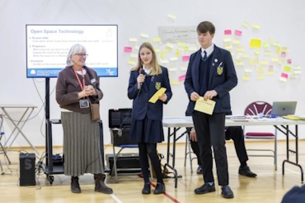 Two students stand alongside a speaker at the Secondary Schools Climate Conference. One of the students is speaking into a microphone.
