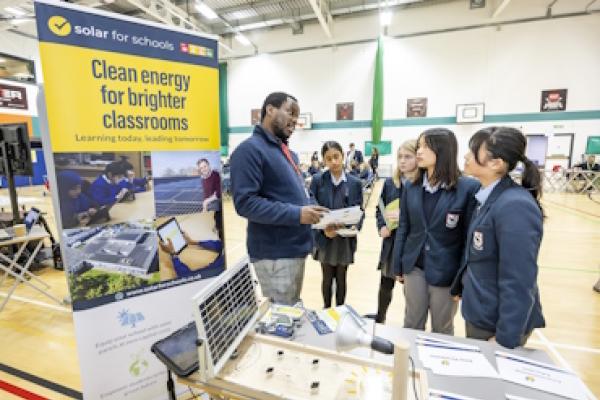 Students talk to a man at a stand from Solar for schools. They stand beside a banner which says "clean energy for brighter classrooms", and there is a model solar panel on the desk.