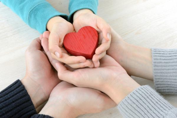 two adults and a child's hands interlocked holding a red wooden heart