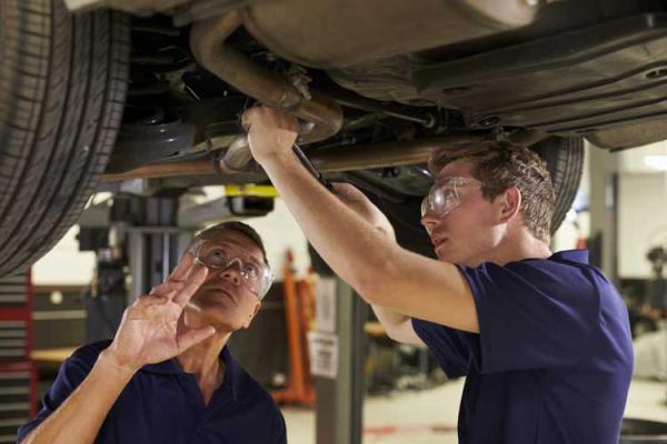 Young person being trained by older person - both looking at the underside of a car.
