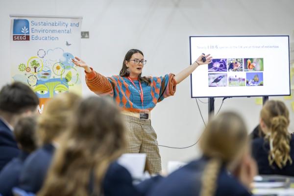 A woman gives a talk to a group of teenagers. She stands with her arms wide, excitedly talking. Behind her is a sign which reads "Sustainability and environmental education".