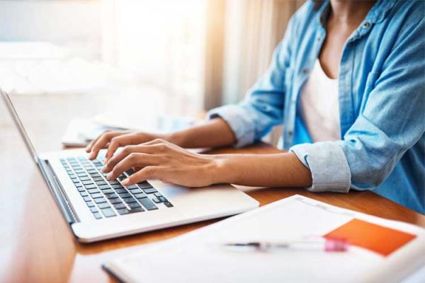Woman wearing blue shirt typing on laptop with A4 pad of paper and pen next to her