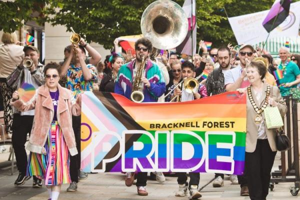 The Bracknell Forest Pride Parade, led by two people holding a Pride banner, followed by a brass band and a crowd of people.