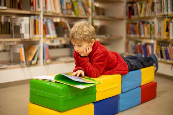 A little boy lies across several stacked soft blocks in a library. A book is open in front of him, which he is reading intently.