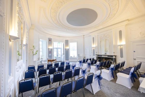 The Haversham Room at South Hill Park, decorated for a wedding or other ceremony. Some of the chairs are covered in white covers with big blue bows.