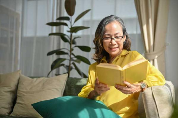 Woman with dark hair and glasses, sitting on a couch, reading a book