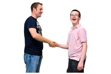 Two smiling people shaking hands, one wearing a dark blue T-shirt and jeans, the other in a pink polo shirt and shorts.
